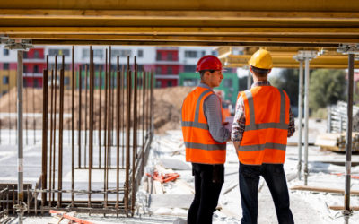 Two civil engineers dressed in orange work vests and helmets discuss the construction process on the building site near the wooden constructions and steel frames